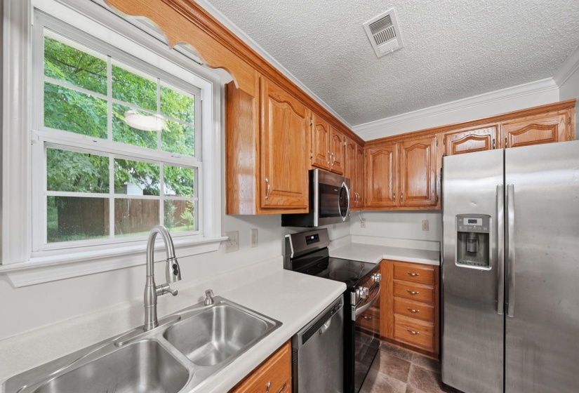 Kitchen featuring wood cabinetry, stainless steel appliances, a double basin sink with a gooseneck faucet, light-toned countertops, and a window overlooking backyard.
