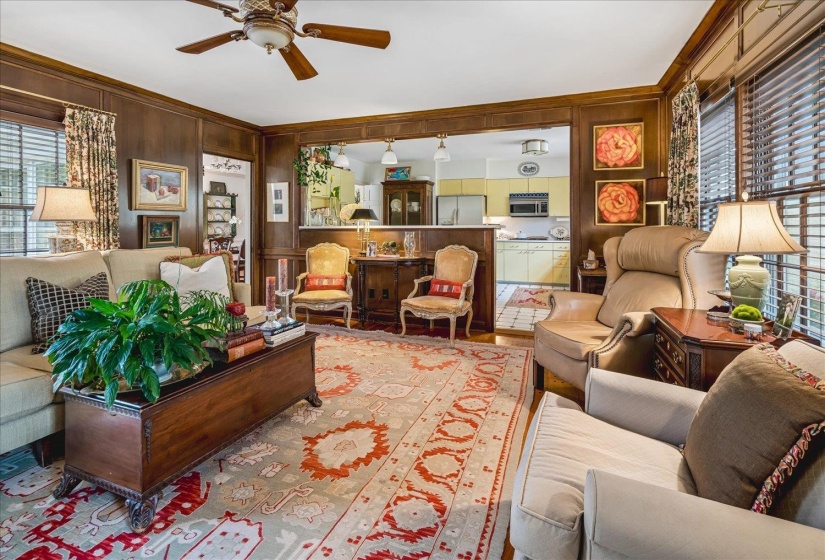 Living area featuring rich wood-paneled walls, a decorative ceiling fan, and a wide cased opening into an adjacent kitchen with light-colored cabinetry