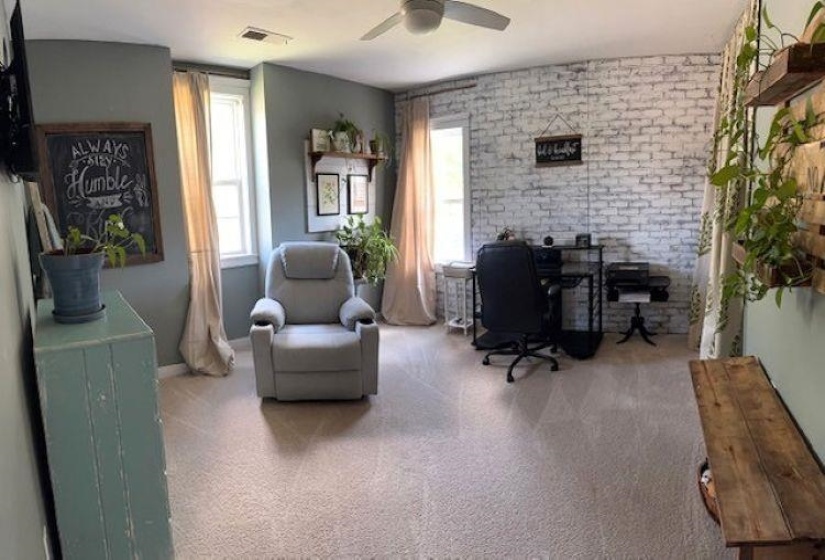 Carpeted room featuring a ceiling fan, multiple windows with natural light, and a white-washed brick accent wall