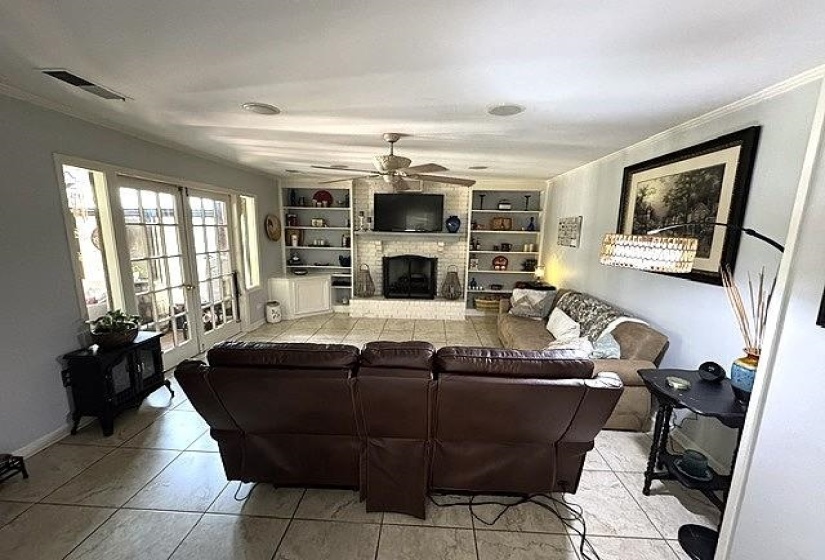 Living area featuring large tile flooring, a fireplace with a white brick surround, built-in shelving, a ceiling fan, and double glass-paneled doors
