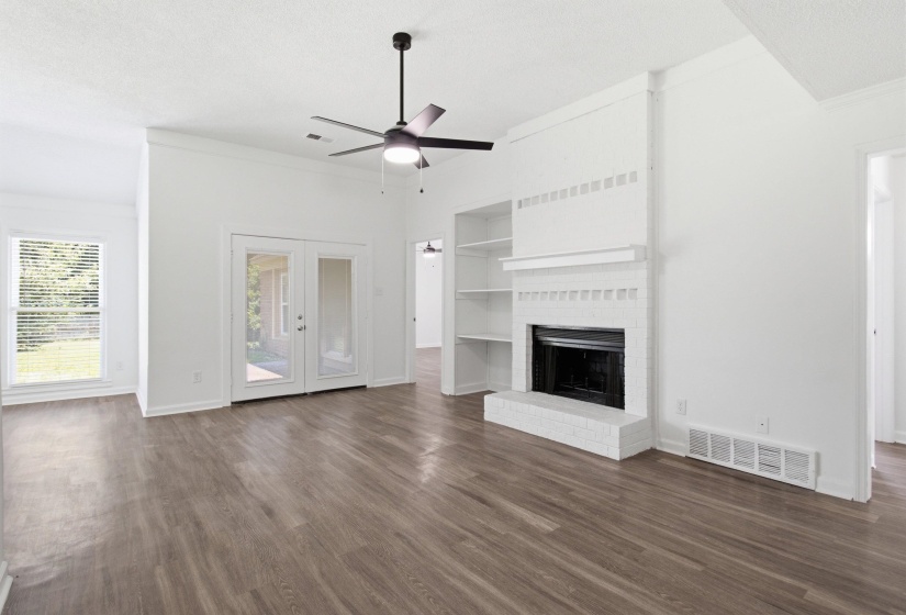 Spacious living area featuring wood-finish flooring, a white brick fireplace with mantel, built-in shelving, a ceiling fan, and French doors