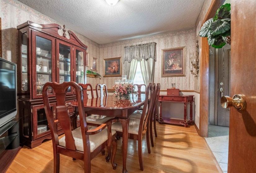 Dining area featuring wood-finish flooring, patterned wallpaper with chair rail molding, and a window with drapes