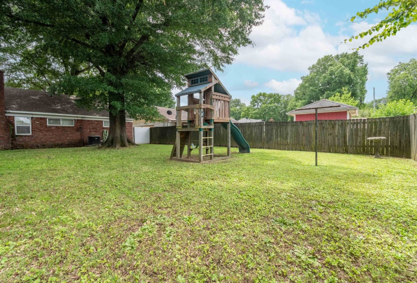 Expansive, fenced backyard featuring a mature shade tree, a wooden play structure with a slide, and a clothesline