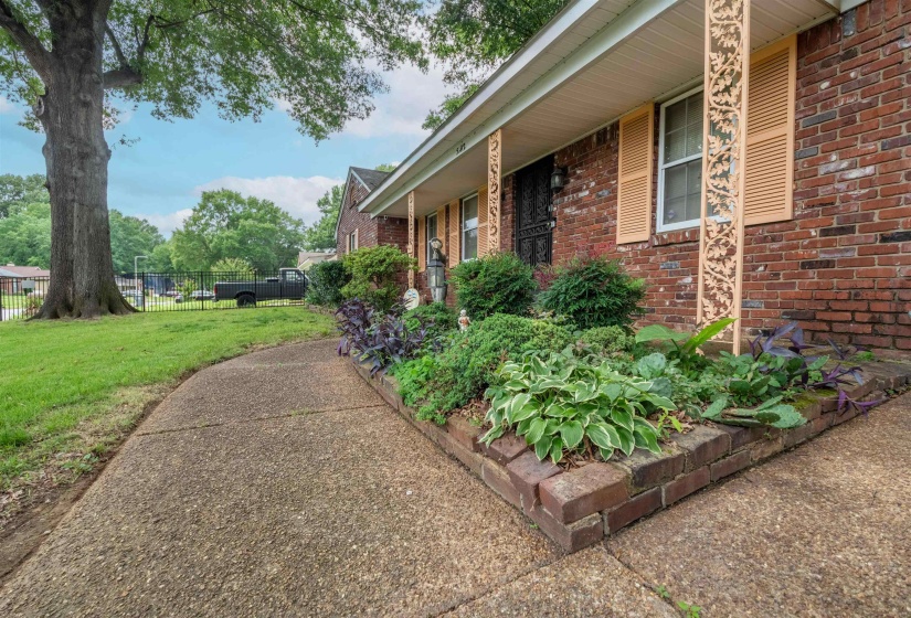 Brick exterior featuring decorative porch columns, louvered window shutters, a concrete walkway, and an elevated garden bed with established plantings