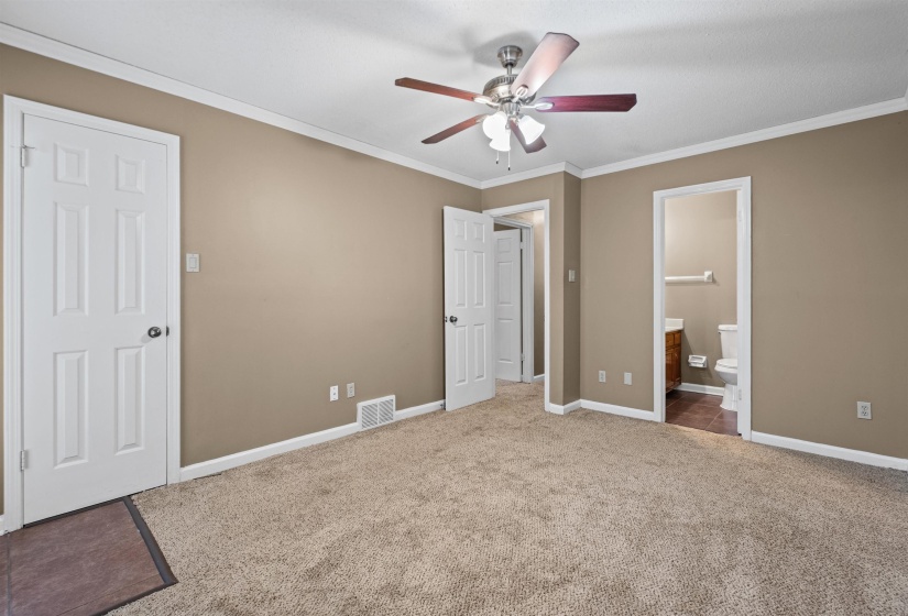 Carpeted bedroom featuring neutral wall tones, white trim, a ceiling fan with light fixture, and an attached bathroom with a wood-finish vanity