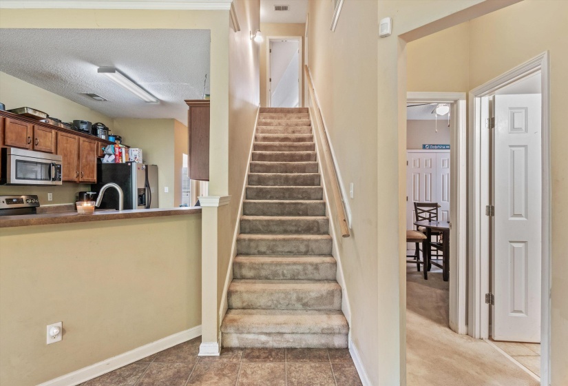 Carpeted staircase with light wood handrail, featuring a partial wall opening to the kitchen