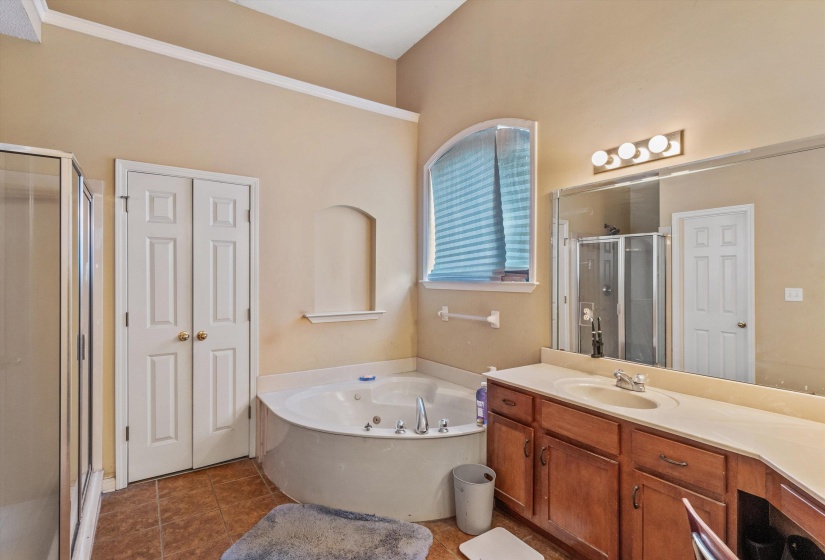 Bathroom featuring a corner jetted tub, arched window with privacy blinds, wood-finish vanity with countertop, and an enclosed shower