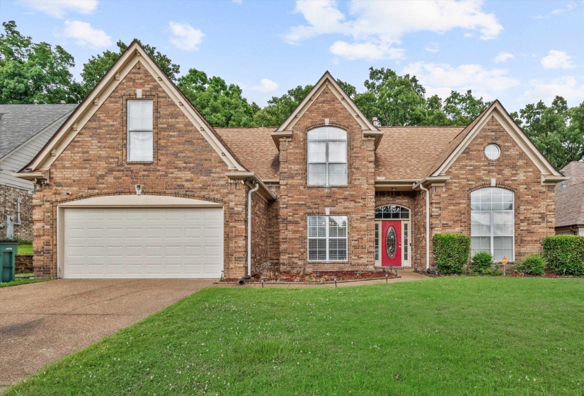 Brick facade residence featuring a prominent red entryway door, an attached two-car garage, multiple gabled rooflines, arched windows, and a landscaped front lawn