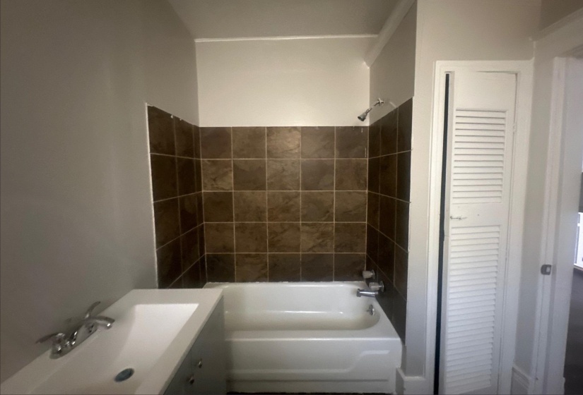 Bathroom featuring a white bathtub, brown tiled shower surround, wall-mounted showerhead, and a white sink with chrome fixtures