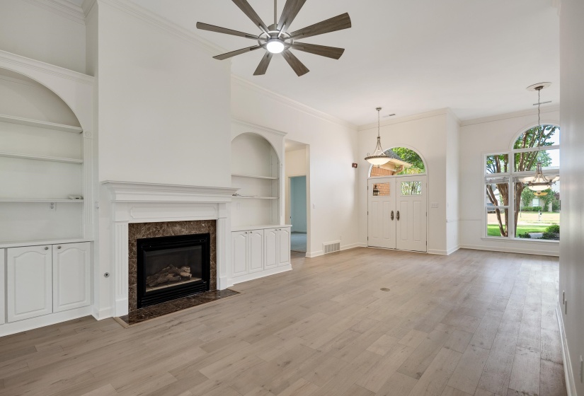 Spacious living area featuring light wood-finish flooring, a central fireplace with a dark stone surround and white mantel, and built-in shelving with lower cabinetry