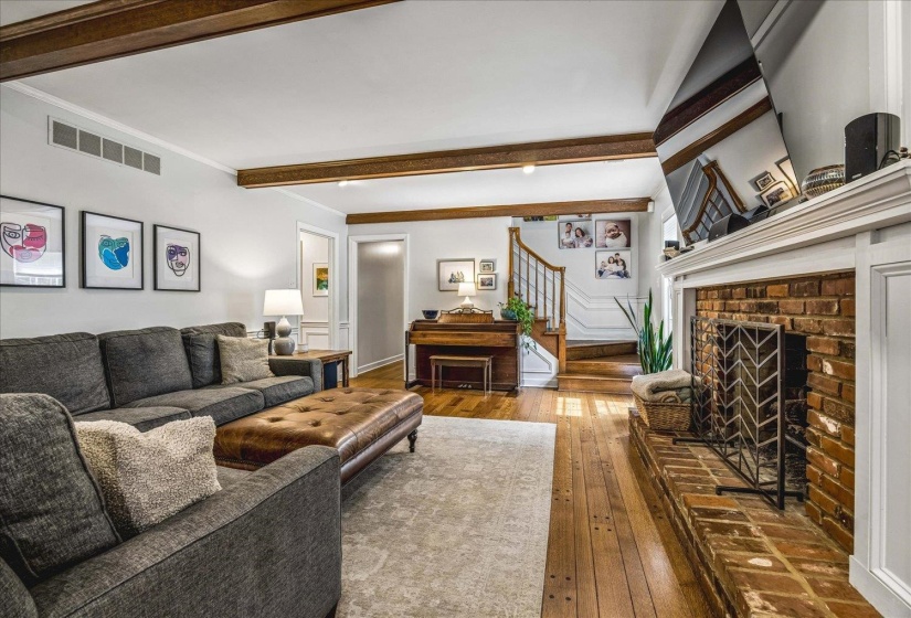 Living area featuring exposed wood beams, brick fireplace with a white mantel, wood-finish flooring, staircase with white risers and wood treads, and white wall paneling
