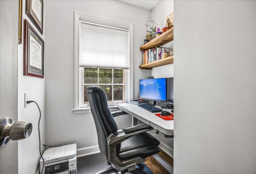 Dedicated workspace featuring a single window with a pleated shade, built-in natural wood shelving, white walls, and dark wood-finish flooring