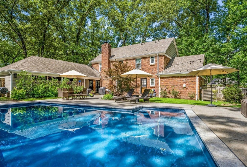 Brick exterior residence featuring a rectangular swimming pool, an expansive concrete patio, a prominent brick chimney, and mature surrounding trees