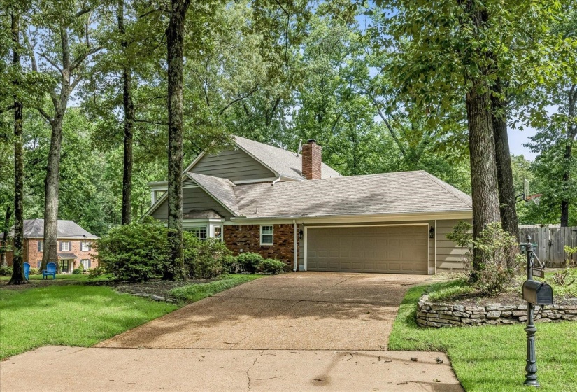Traditional-style residence featuring a brick and siding exterior, a two-car garage, an exposed aggregate driveway, a stone-lined garden bed, and mature trees providing natural shade