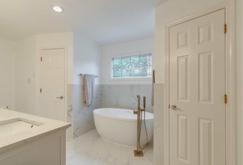 Freestanding soaking tub featuring a floor-mounted faucet with hand shower, marble-finish wall and floor tiling, and a decorative stained glass window
