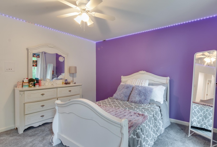 Bedroom featuring a ceiling fan, purple accent wall, carpeted flooring, and decorative perimeter lighting