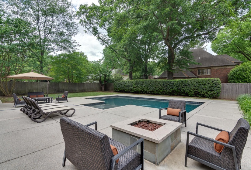 Inviting outdoor area featuring a rectangular swimming pool with a surrounding paved deck, a stone-clad fire pit, and mature perimeter trees