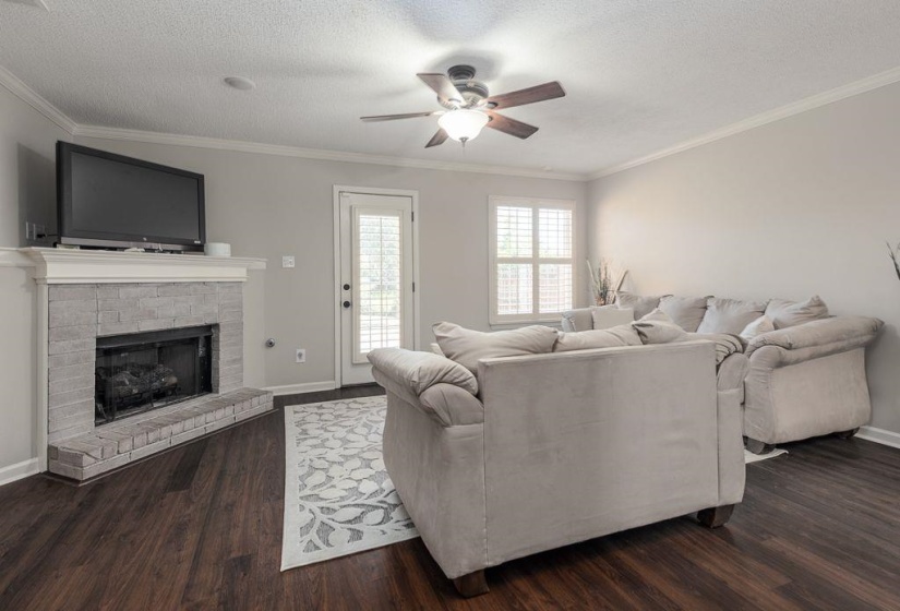 Living area featuring dark wood-finish flooring, a light gray brick fireplace with a white mantel, and a ceiling fan with integrated lighting