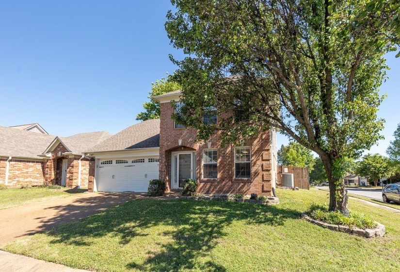 Brick exterior featuring a two-car garage, mature trees, and a landscaped front yard