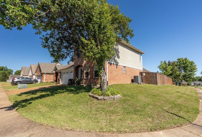 Two-story brick and siding residence featuring an attached garage, a mature tree with a stone-edged planting bed, a private wood fence, and a spacious front lawn