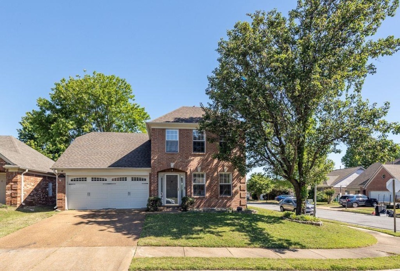 Brick facade two-story residence featuring an attached two-car garage, pitched shingle roof, and a mature shade tree
