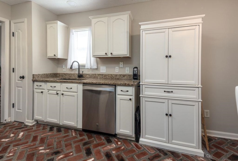 Kitchen featuring brick-finish flooring, white cabinetry with dark hardware, granite countertops, a stainless steel dishwasher, and a window with white drapes