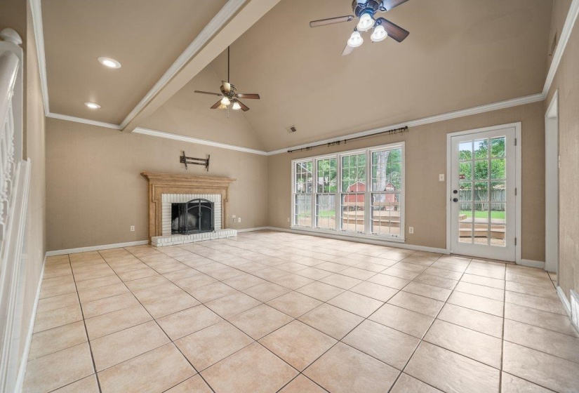 Spacious living area featuring neutral-toned tile flooring, a fireplace with a white brick surround and carved wood mantel, and multiple ceiling fans