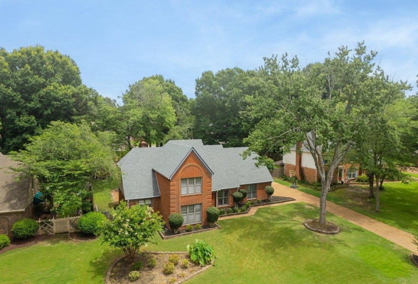 Brick facade home featuring a gabled roofline, mature trees, manicured landscaping, and a broad driveway