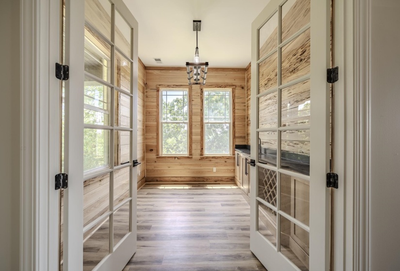 Double French doors opening to an interior room with wood plank walls, light wood-finish flooring, two large windows, and built-in cabinetry with a dark countertop and wine rack