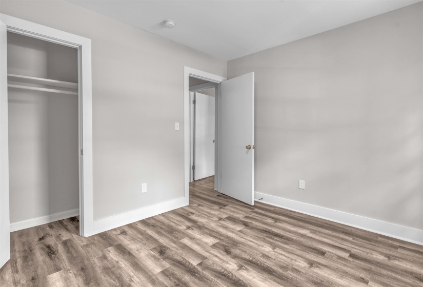Spacious room featuring wood-finish flooring, light gray wall color, white trim, a built-in closet with shelving and hanging rod, and a white interior door with brass hardware