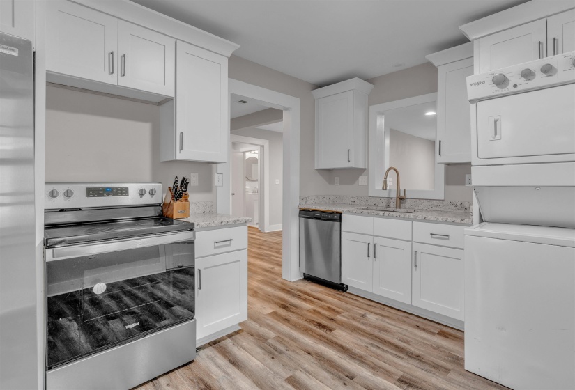 Bright kitchen featuring white shaker-style cabinetry, light-toned granite countertops, wood-finish flooring, stainless steel appliances, and a stacked laundry unit