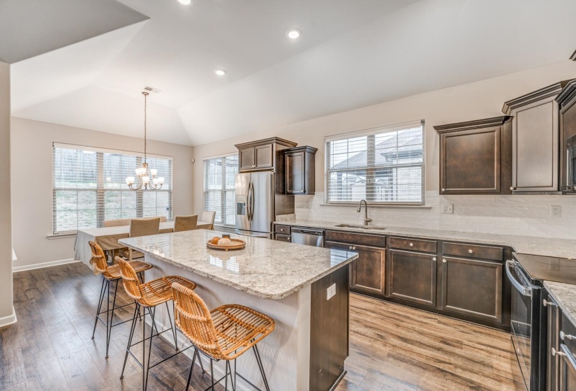 Kitchen featuring dark wood finish cabinetry, backsplash, stainless steel appliances, light stone countertops, and vaulted ceiling