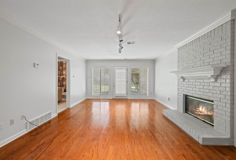Expansive room featuring rich wood-finish flooring, a painted brick fireplace with a white mantel, and white plantation shutters on double French doors