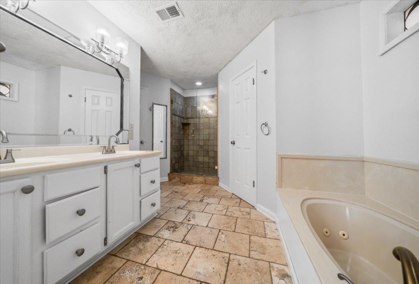 Spacious bathroom featuring natural stone tile flooring, a double vanity with white cabinetry, a built-in jetted tub, and a separate walk-in shower with tiled walls