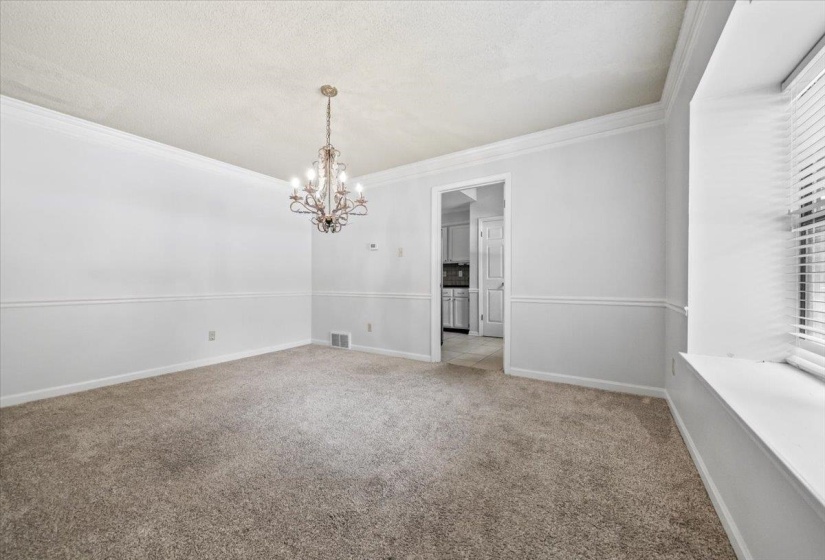 Spacious room featuring neutral carpeting, a decorative chandelier, crown molding, chair rail detailing, and a large window with blinds