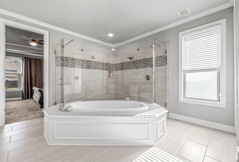 Expansive bathroom featuring a corner soaking tub with paneling, a large glass-enclosed shower with dual shower heads, decorative tile accents, light wood-finish flooring, and a window with blinds