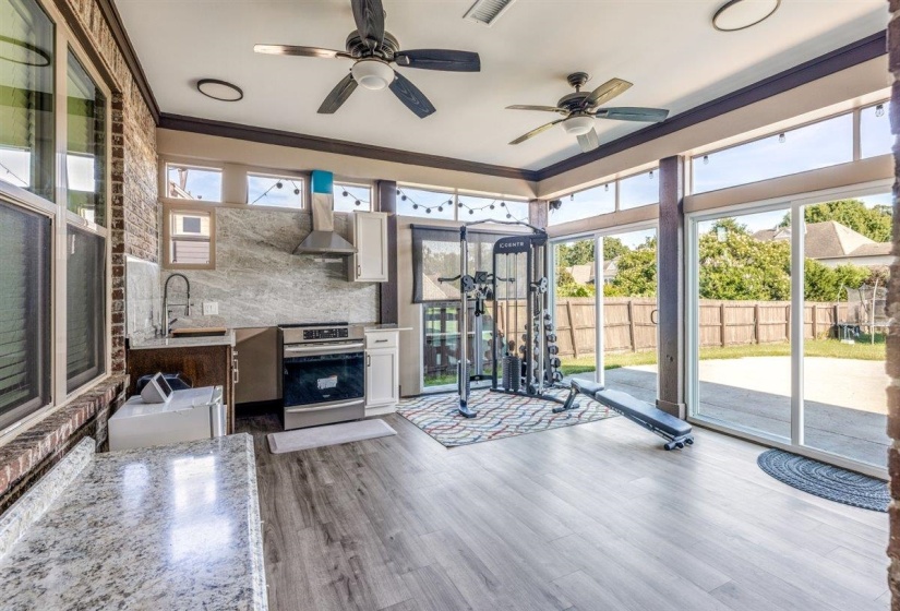 Spacious sunroom featuring wood-finish flooring, two ceiling fans, a kitchenette with stainless steel appliances and a stone backsplash, and expansive sliding glass doors