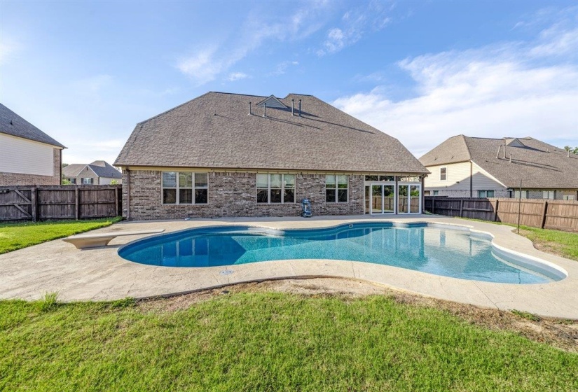 Curved inground pool with concrete surround, brick exterior, and a privacy fence