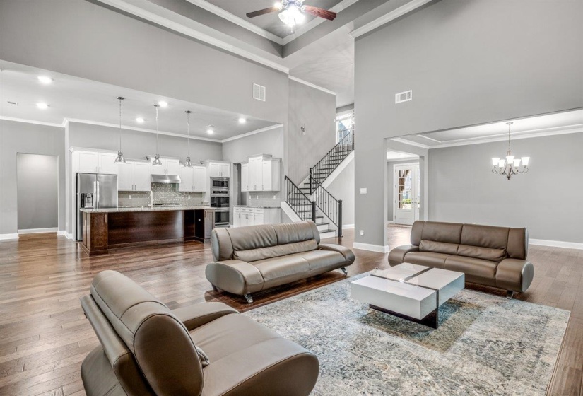 Spacious open-concept interior featuring wood-finish flooring, a two-story ceiling, and a kitchen with white cabinetry, stainless steel appliances, and an island with pendant lighting