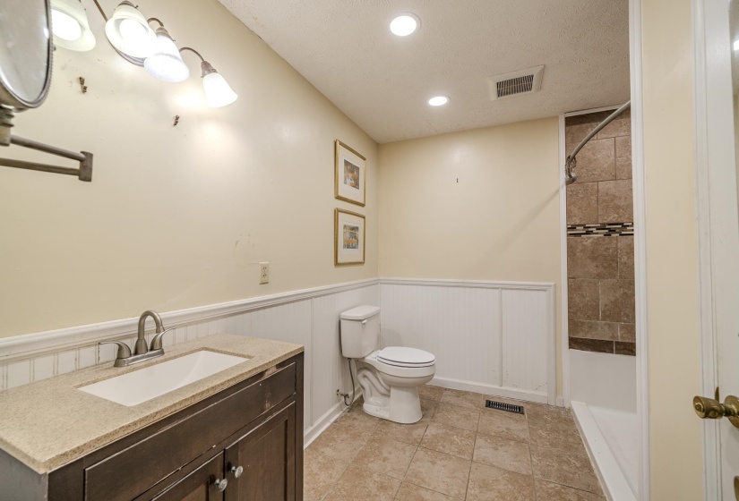 Bathroom featuring a dark wood-finish vanity with an integrated sink, a light-toned countertop, beadboard wainscoting, and a tiled shower with decorative accent banding