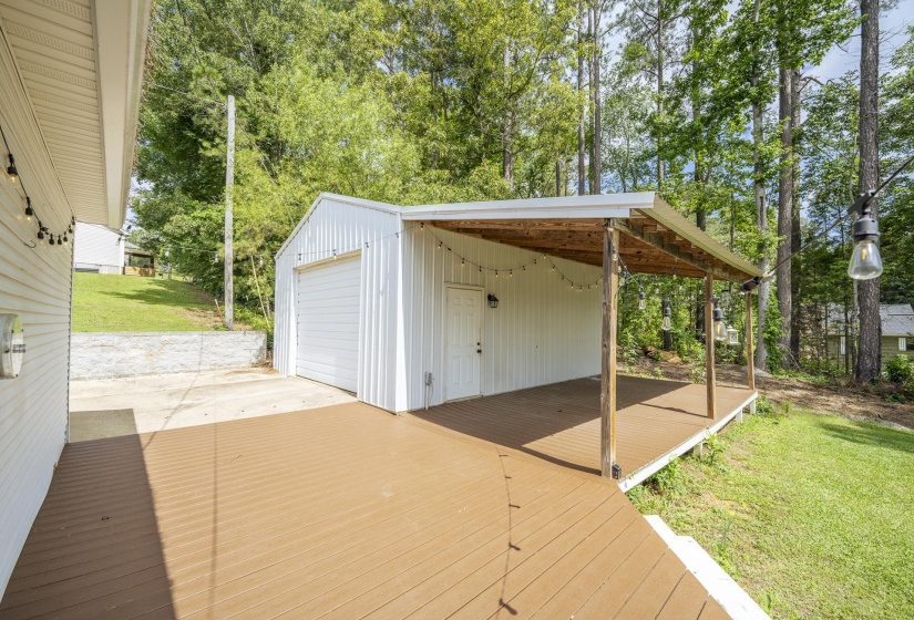 Detached white metal garage featuring a roll-up door and an attached covered patio with wood-finish decking and exposed wood support posts