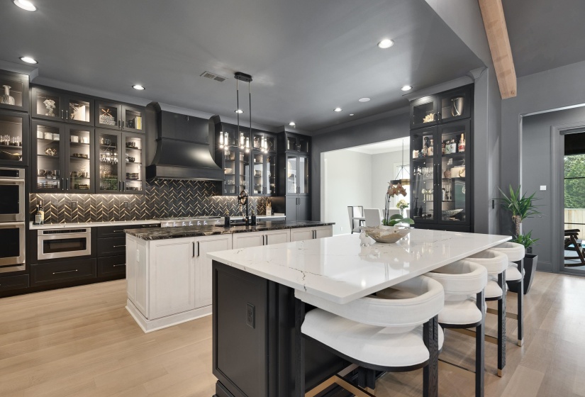 Gourmet kitchen featuring a large quartz island, light wood-finish flooring, built-in stainless steel appliances, herringbone backsplash, and illuminated glass-front cabinetry