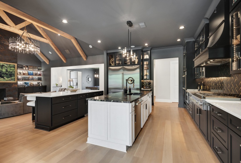 Gourmet kitchen and living area featuring wood-finish flooring, exposed wood ceiling beams, two kitchen islands with stone countertops, stainless steel appliances, and built-in cabinetry