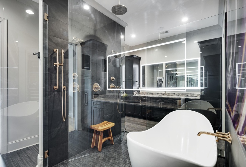 Spacious bathroom featuring a freestanding soaking tub with brass fixtures, a glass-enclosed shower with multiple showerheads, dark vanity cabinetry with integrated sinks, and illuminated rectangular mirrors