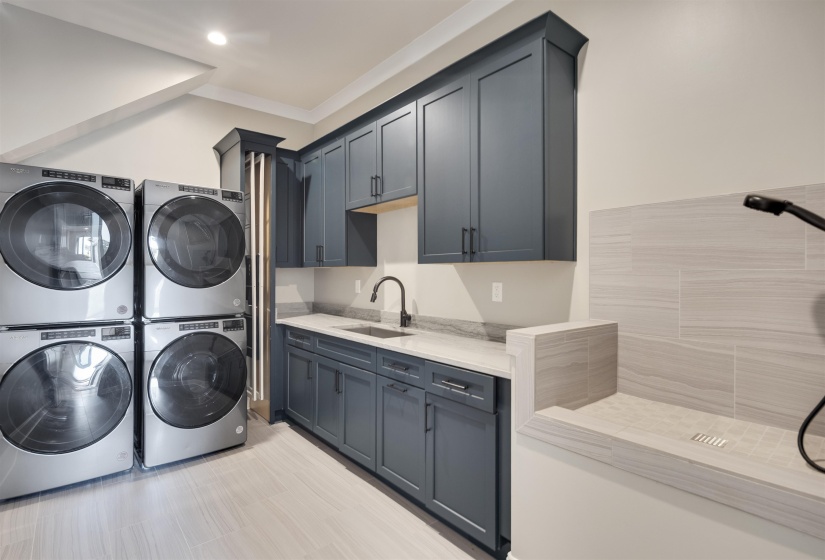 Contemporary laundry room featuring slate blue cabinetry, a built-in sink with a matte black faucet, light stone countertops, wood-finish flooring, and a dedicated wash station with tiled walls