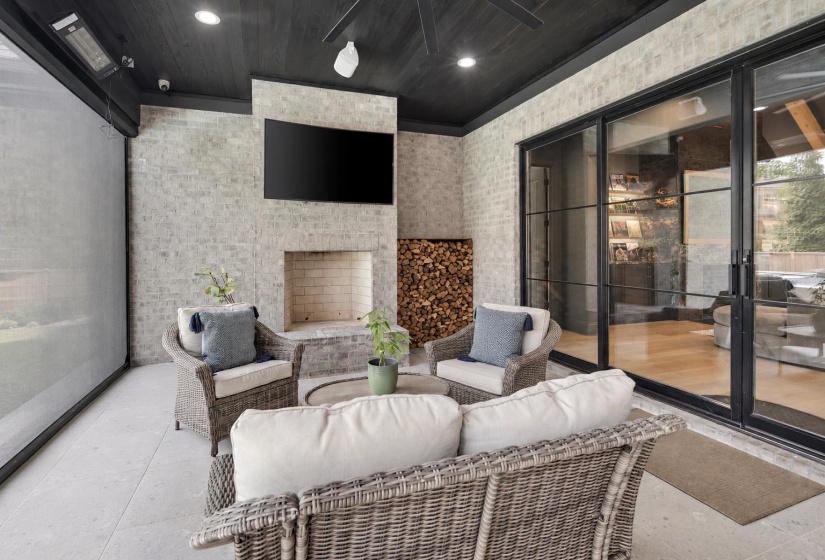 Covered outdoor living space featuring light-colored brick walls, a built-in fireplace, and a dark wood-finish ceiling with recessed lighting