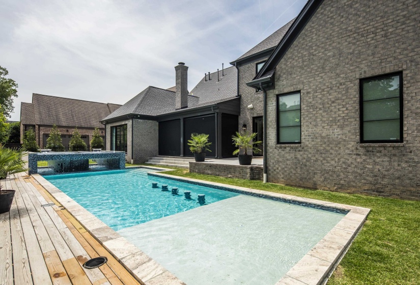 Pool with an integrated sun shelf and water feature, complemented by a wood-finish deck and surrounding stone coping
