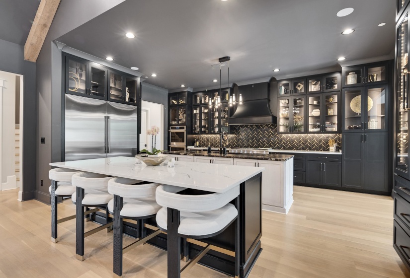 Gourmet kitchen featuring a large central island with seating, stainless steel double refrigerator, integrated hood vent, herringbone tile backsplash, and light wood-finish flooring