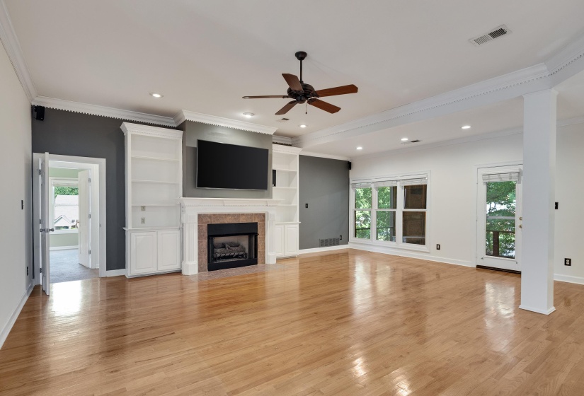 Spacious living area featuring polished wood-finish flooring, a central fireplace with stone surround and white mantel, integrated built-in shelving, a ceiling fan, and multiple windows offering natural light