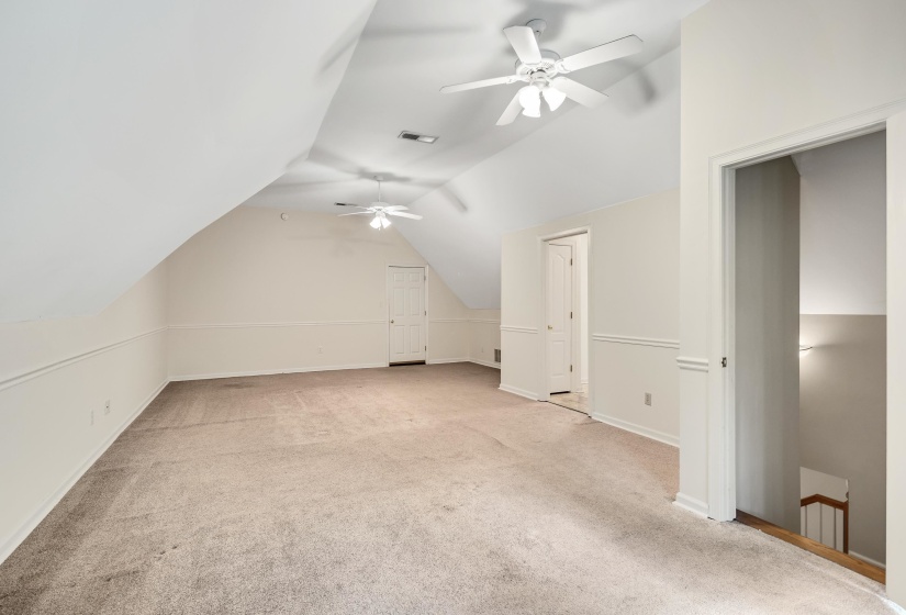 Spacious carpeted room featuring angled ceilings, two ceiling fans, chair rail molding, and multiple white paneled doors